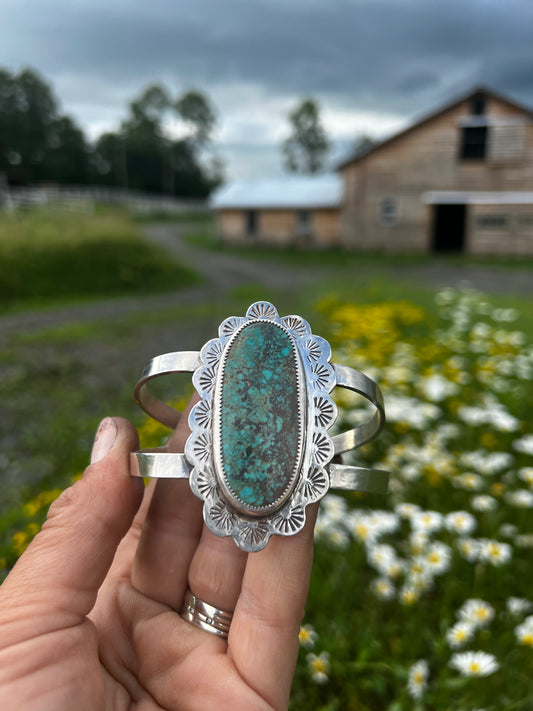 A sterling silver cuff bracelet by Red Pony Brand with a large Royston Turquoise stone held in front of wildflowers and aa wood sided horse barn