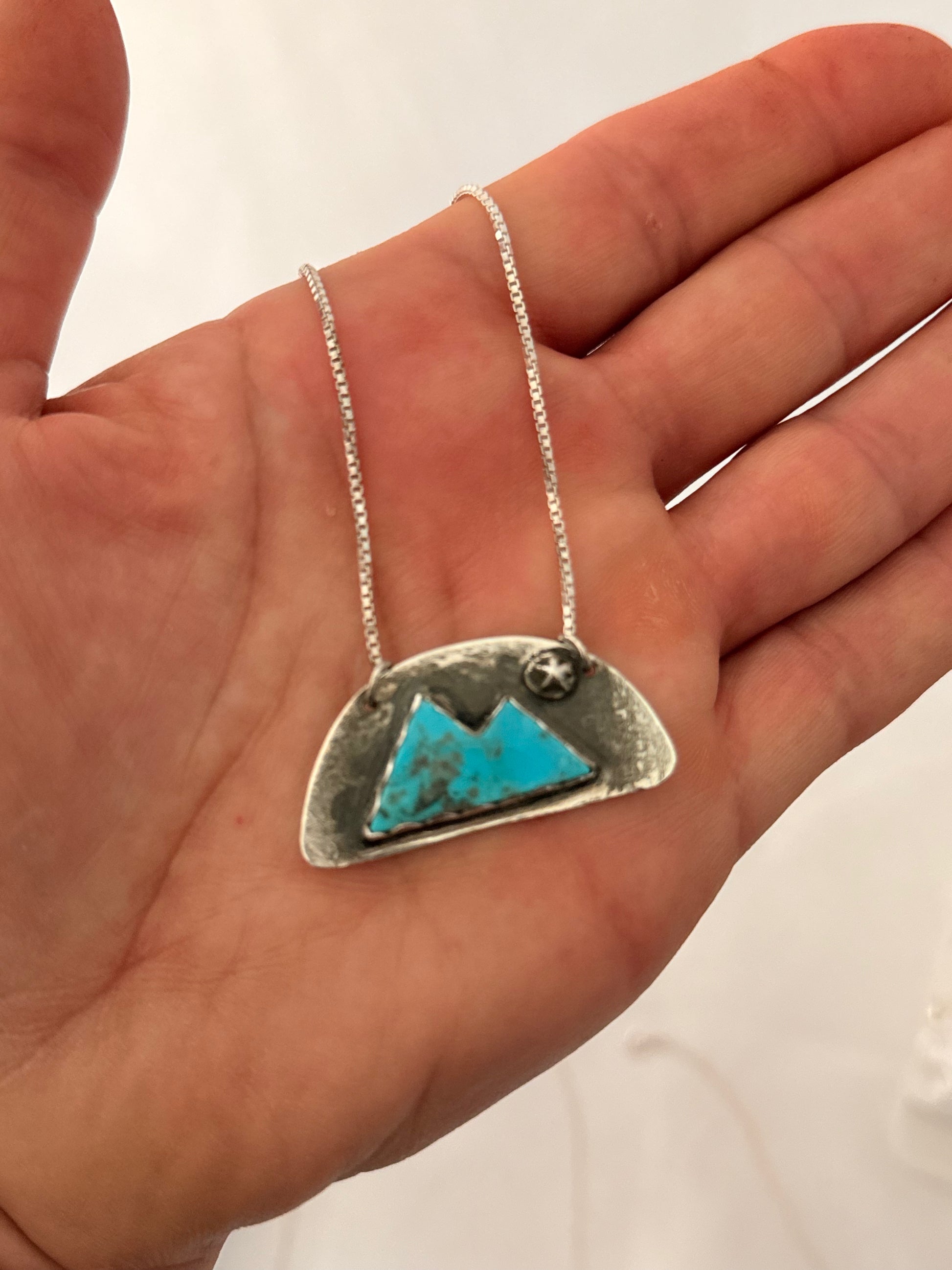 Red Pony Brand silver half circle pendant with a turquoise stone cut in the shape of a mountain held in a hand with a white background